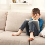 Gutt sitter i sofa, holder seg for øynene, har et nettbrett i fanget. Foto: Shutterstock.