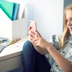 Foto: Shutterstock / Monkey Business Images. Bildet viser ei tenåring ved pulten sin hjemme. Hun har PC og blyanter på bordet. I hånden holder hun en telefon.