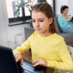 Foto: Shutterstock / Syda Productions. Bildet viser ei jente som skriver på tastaturet til en datamaskin hjemme i stua, moren sitter og leser en bok i sofaen i bakgrunnen.