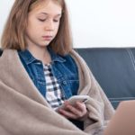 Foto: Shutterstock / PV productions. Bildet viser ei jente i sofaen, med teppe rundt seg, hun ser på telefon og har datamaskin på fanget.